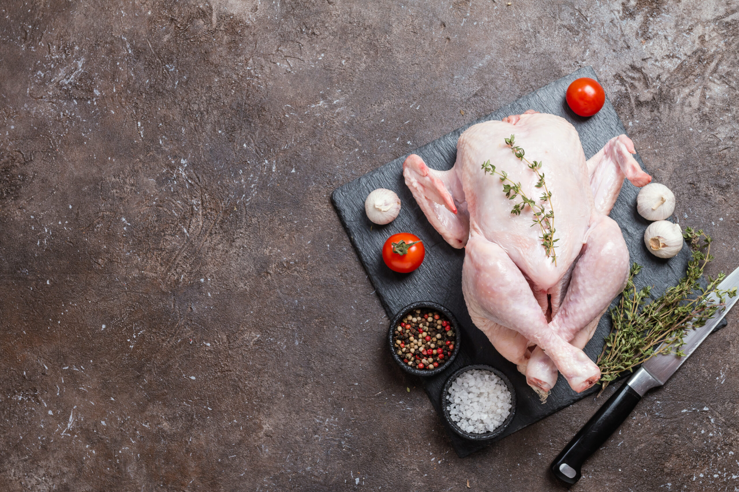 Raw whole chicken on a black slate cutting board, surrounded by fresh herbs, cherry tomatoes, garlic, and bowls of mixed peppercorns and sea salt, with a chef's knife nearby.