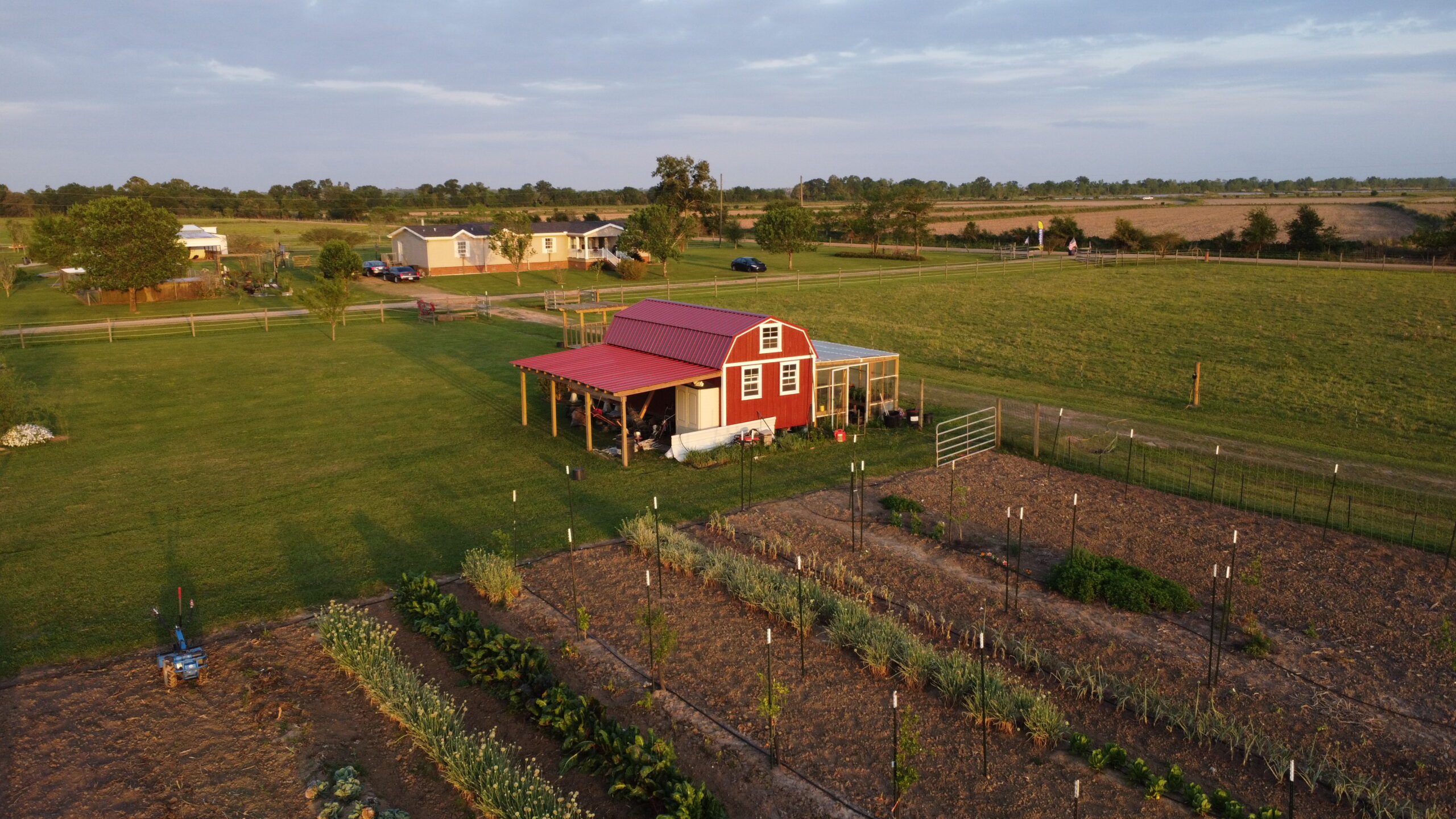 Aerial view of a red barn-style shed surrounded by lush green lawns and a vegetable garden featuring neatly arranged plants, with additional houses and farmland in the background.