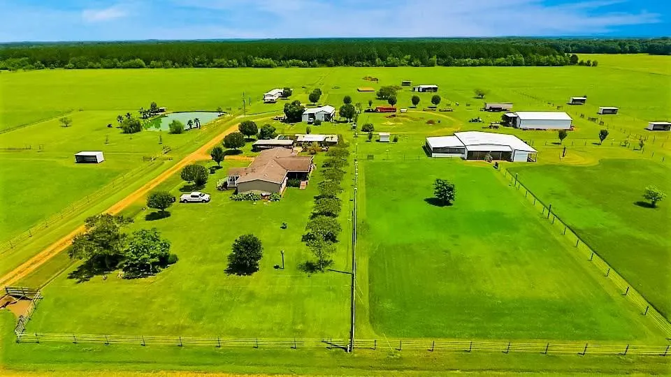 Aerial view of a spacious rural property featuring lush green fields, a large house, several outbuildings, and a pond, surrounded by trees and fencing. Ideal for farming or outdoor activities.
