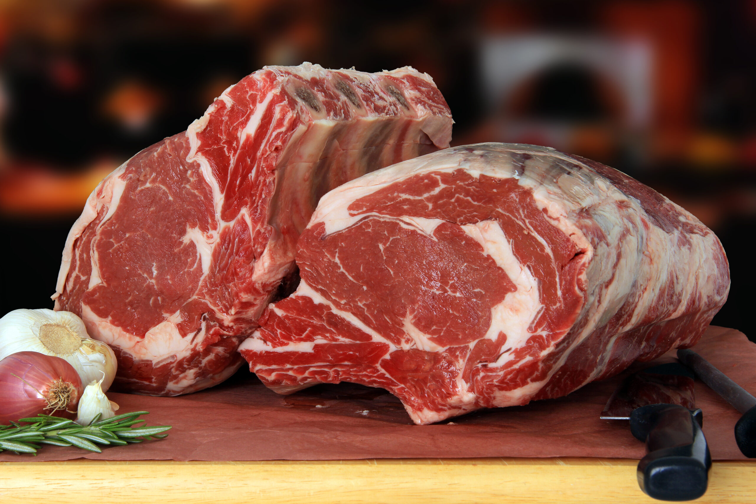 Two large cuts of ribeye beef, showcasing marbling and rich red color, accompanied by garlic, shallots, and fresh rosemary on a wooden surface, perfect for gourmet cooking.